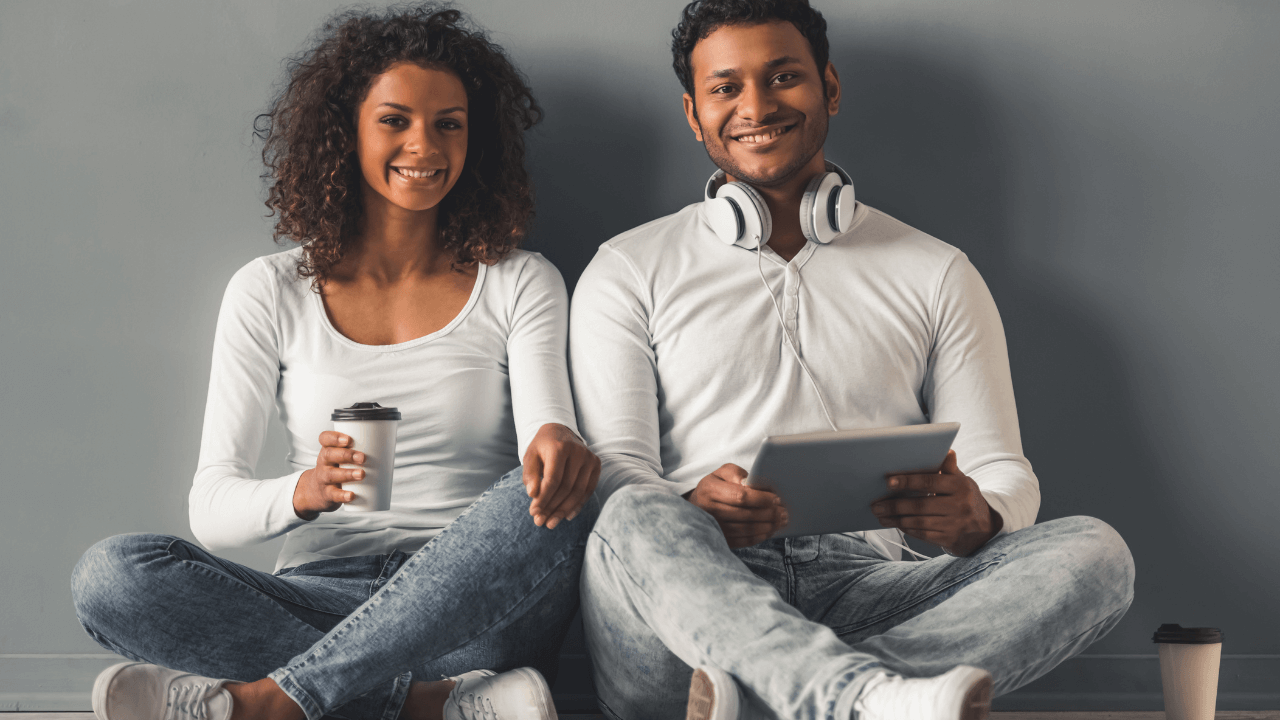 Un couple assis face à une caméra, l'homme tient une tablette tactile tandis que la femme tient un gobelet à café